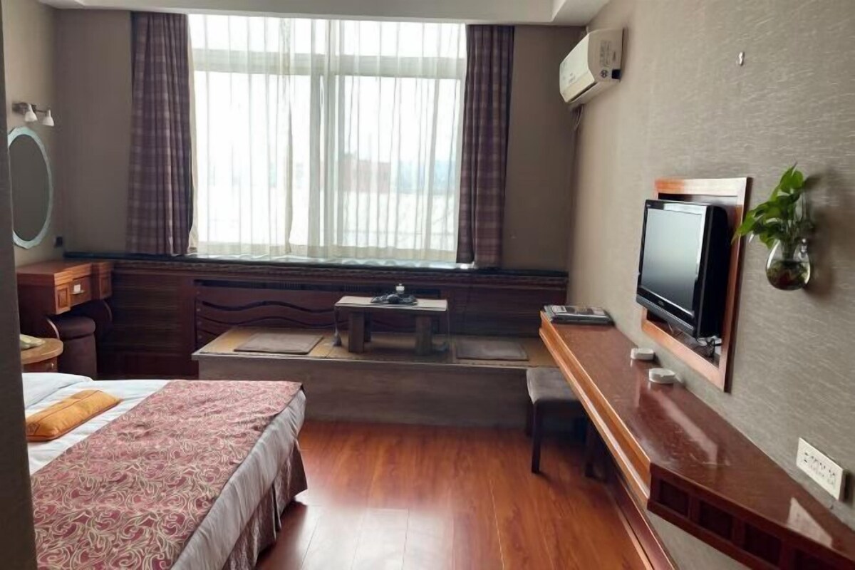 Quarto de hotel com cama, escrivaninha com televisão, mesinha e janela com cortinas.
