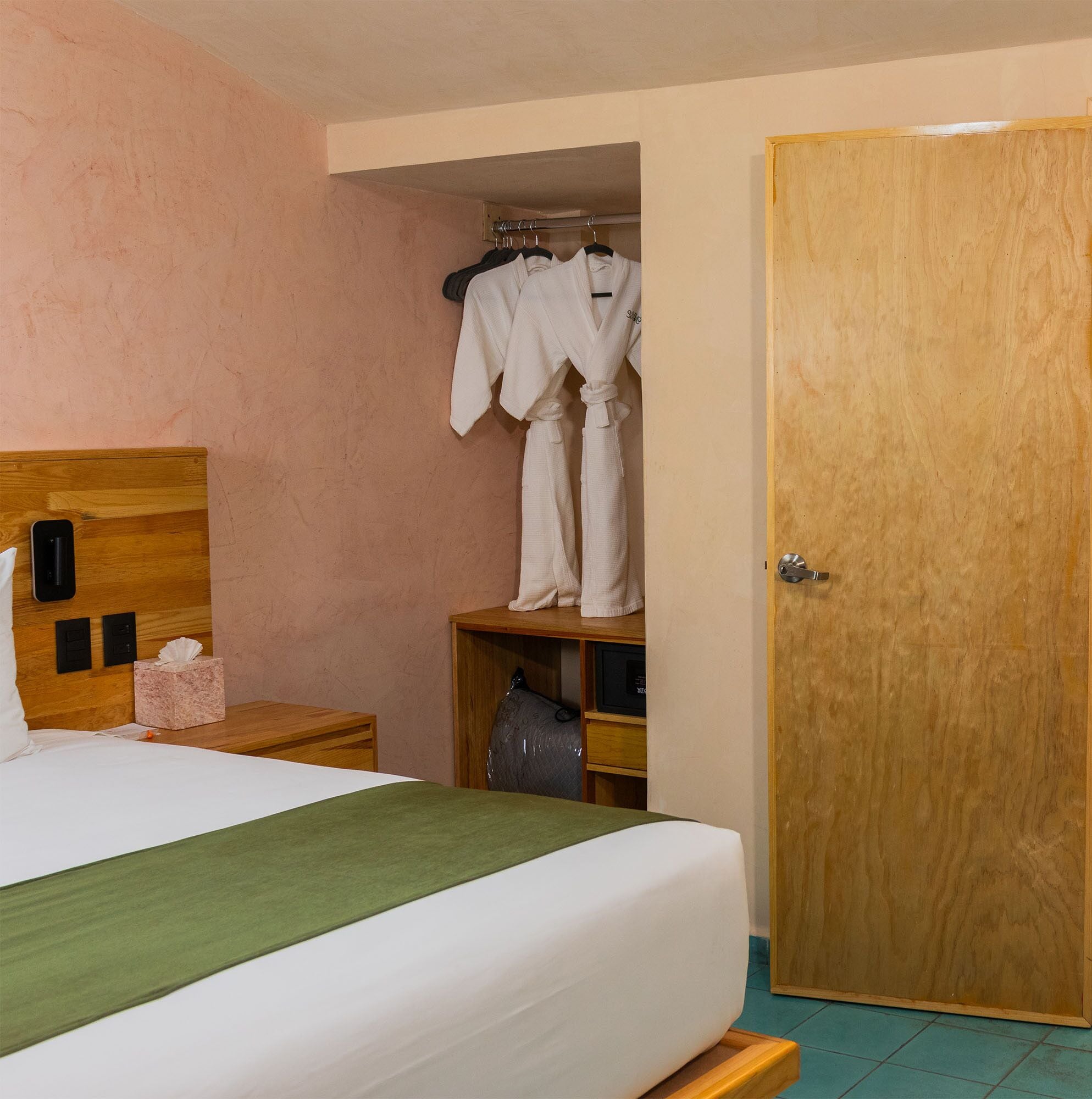 A hotel room with a wooden headboard, a bed with a green bedspread, a bedside table with a tissue box, a wooden nightstand, a wooden wardrobe with a robe hanging, and a wooden door.