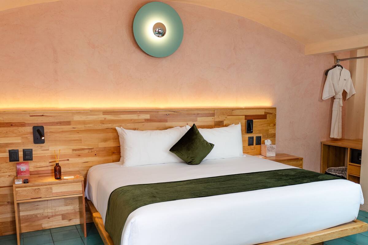 A hotel room with a wooden headboard, a bed with white bedding and a green throw, a bedside table with a bottle and a tissue box, and a robe hanging on a rack.