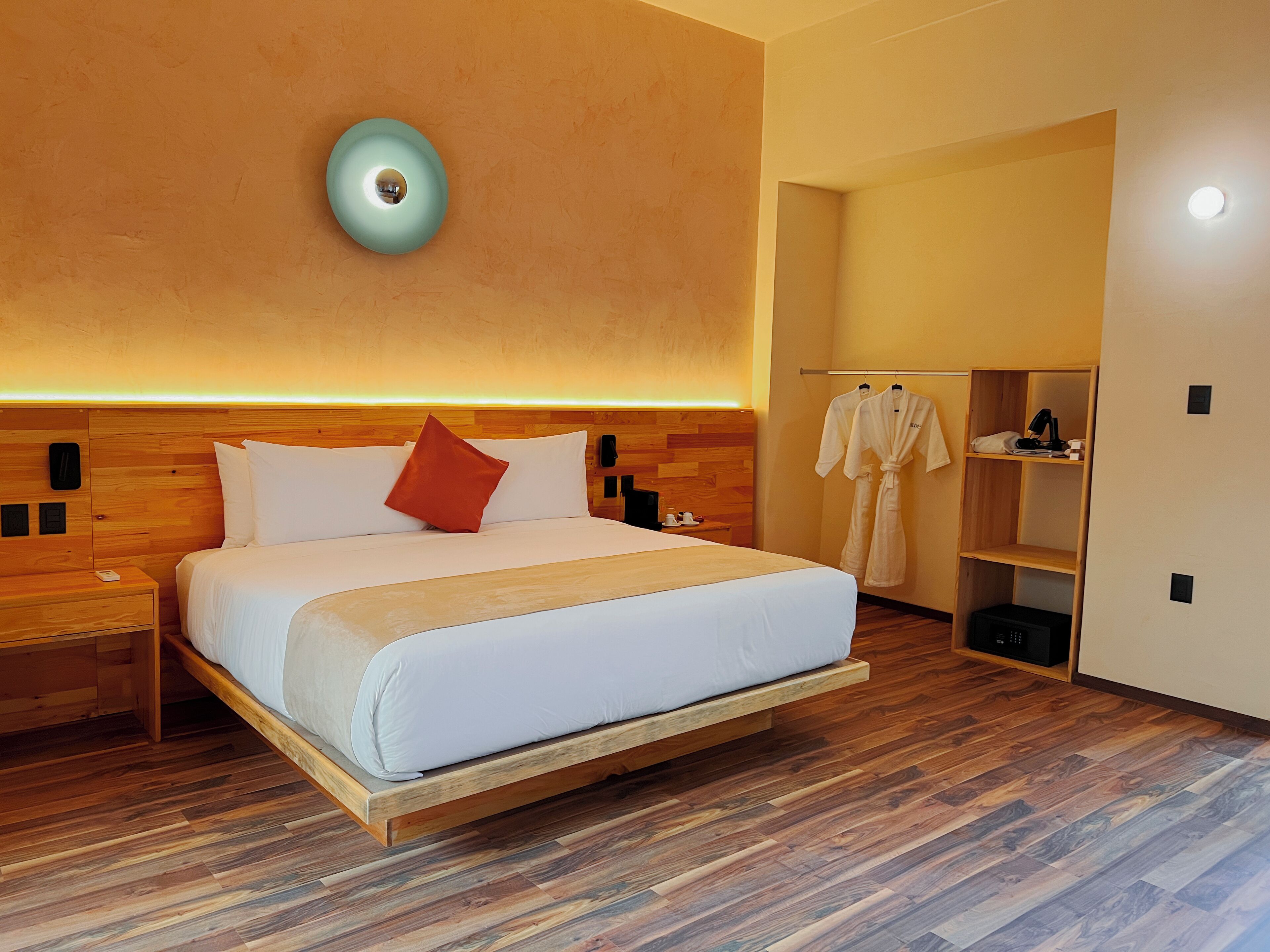 Quarto de hotel moderno com cabeceira de madeira, uma cama grande com roupa de cama branca, um travesseiro vermelho e uma mesa de cabeceira com abajur.