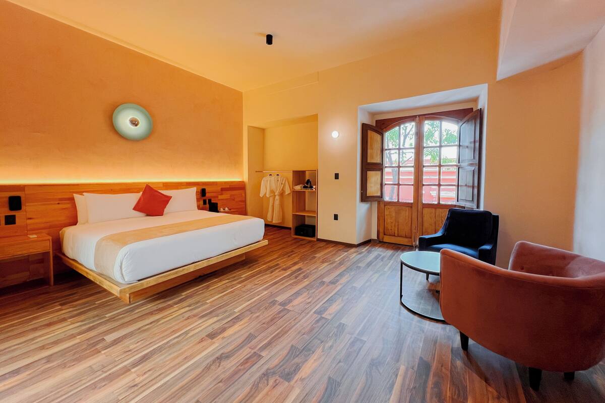 A hotel room with a wooden bed, a round wall clock, a seating area with a chair, and a large window with a view.