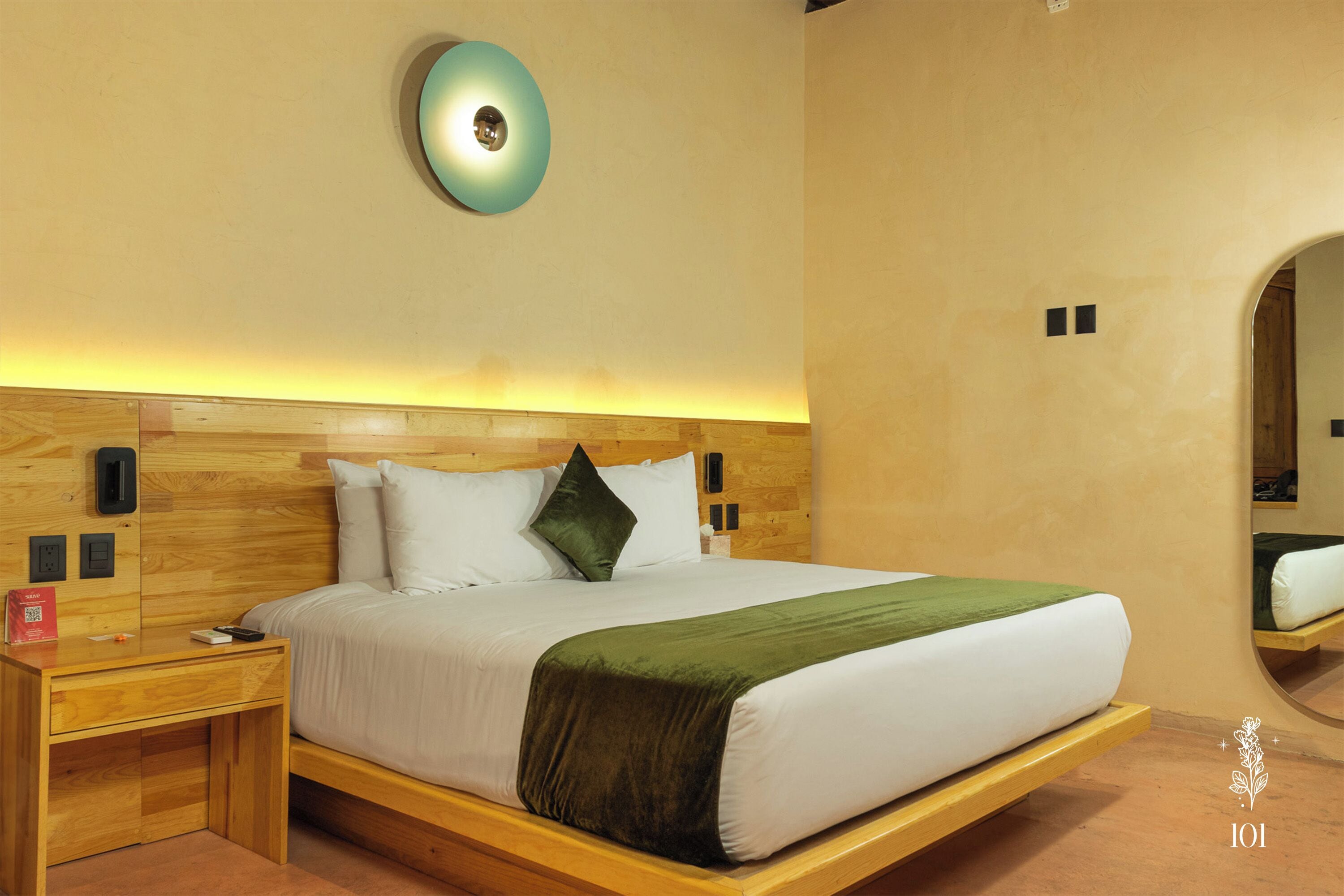 Una habitación de hotel moderna con cabecera de madera, una manta verde y una mesita de noche con un objeto rojo.