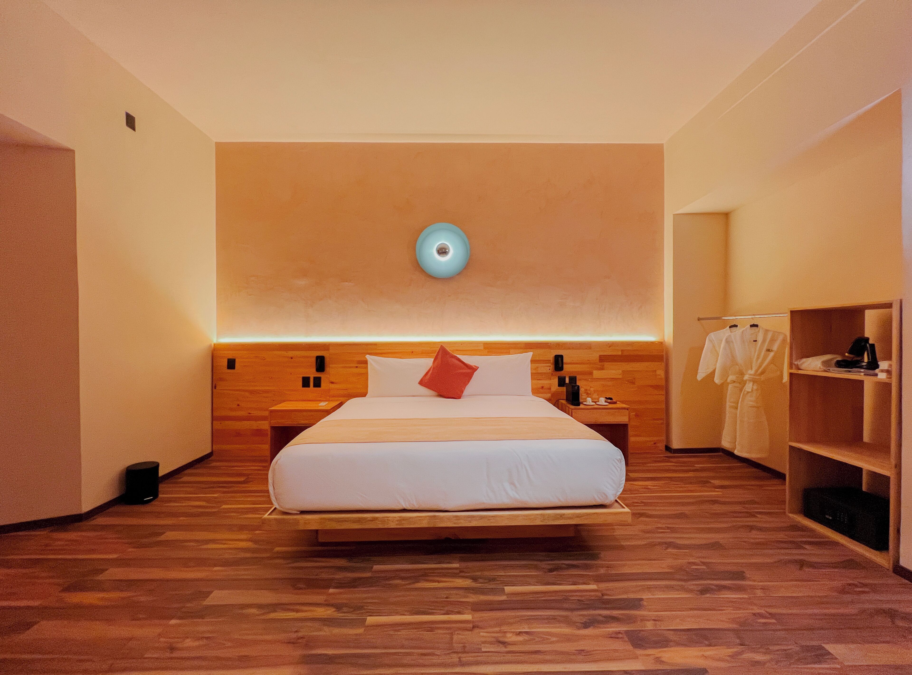 A modern hotel room with a wooden bed, a nightstand, a wall-mounted light, and a robe rack.