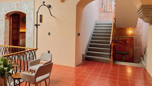 Un interior amplio con una escalera que lleva a un nivel superior, una zona de estar con una silla y una mesa, y una pared decorativa con un cuadro.