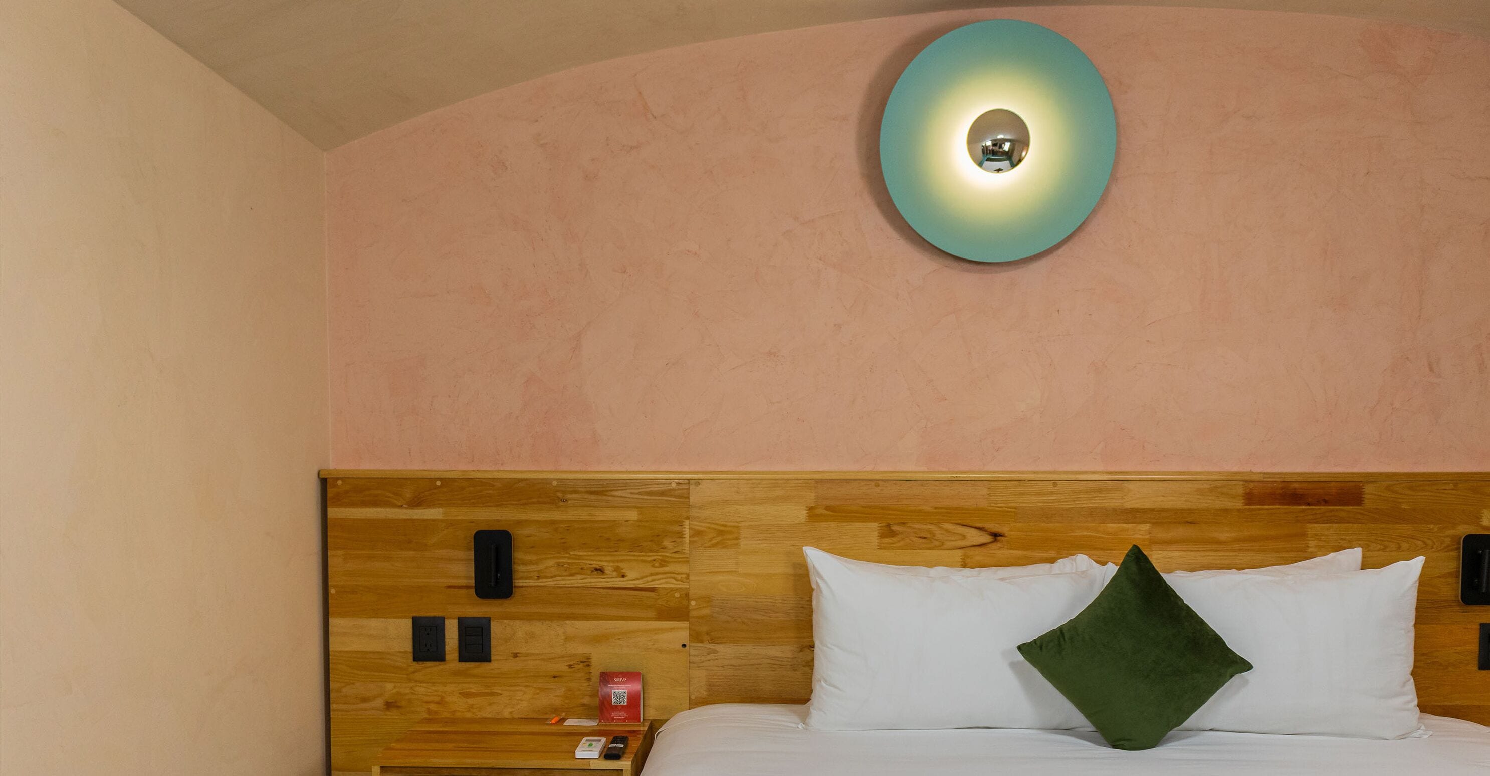 Une chambre d’hôtel avec une tête de lit en bois, une table de chevet avec un téléphone et un objet rouge, et un oreiller vert sur le lit.
