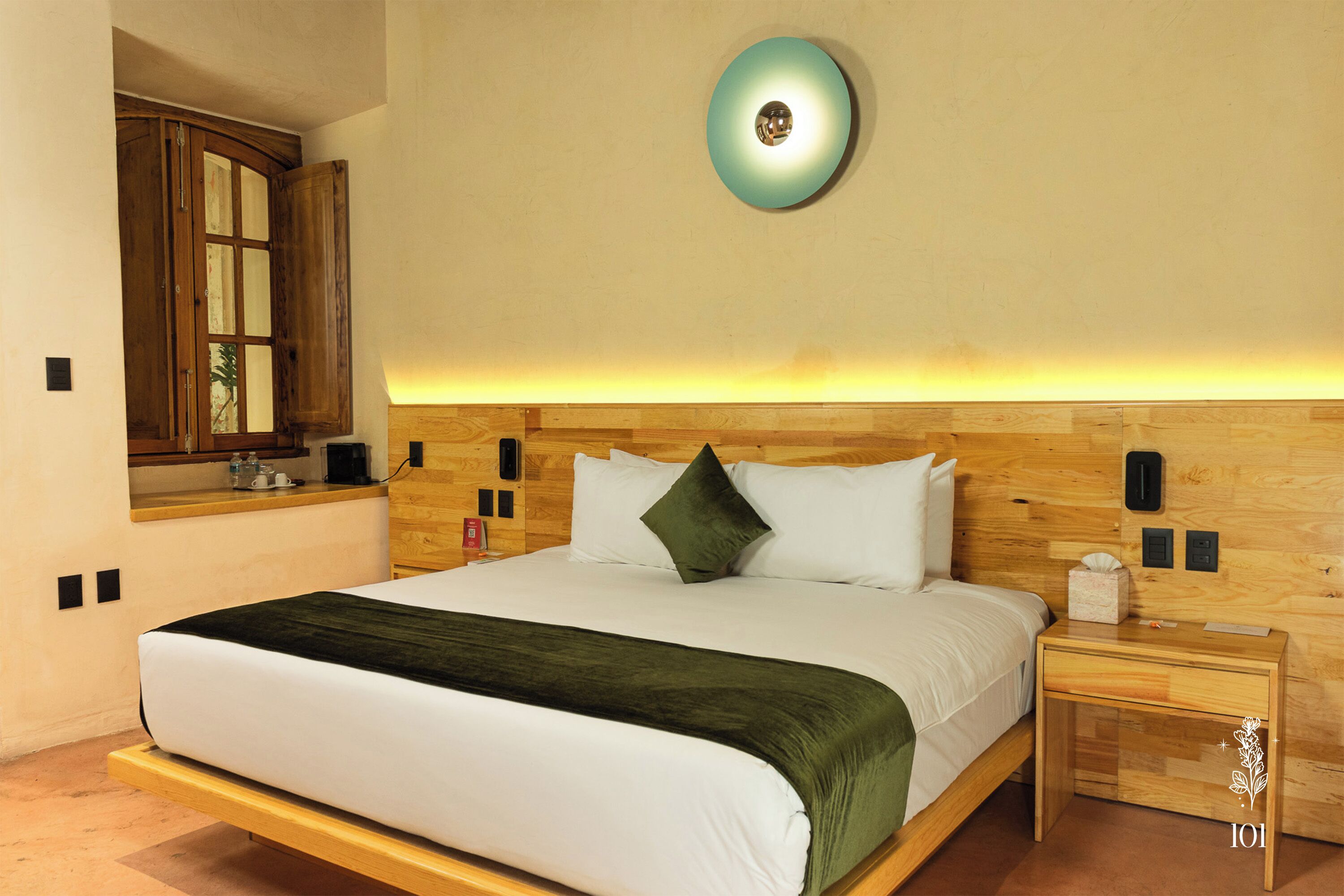 Un dormitorio con cama, mesita de noche, tablero lateral, cabecero de madera, luz adosada a la pared y ventana con cortinas.