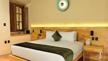 Un dormitorio con cama, mesita de noche, tablero lateral, cabecero de madera, luz adosada a la pared y ventana con cortinas.