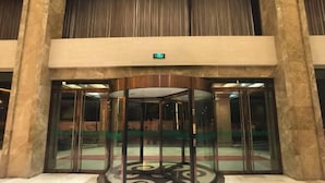 Een draaideur met glazen panelen en een centraal rond ontwerp, die leidt naar een lobby met marmeren vloeren en een versierde zuil.