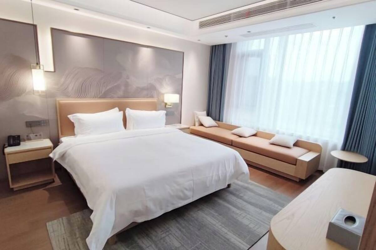 A modern hotel room with a large bed, a sofa, a desk, and a window with curtains.