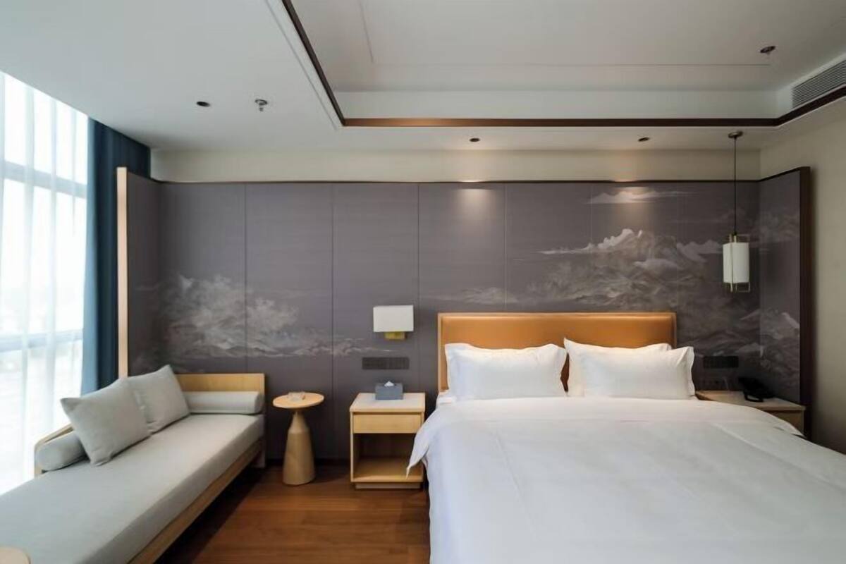 A modern hotel room with a large bed, a sofa, a nightstand, and a wall-mounted lamp.