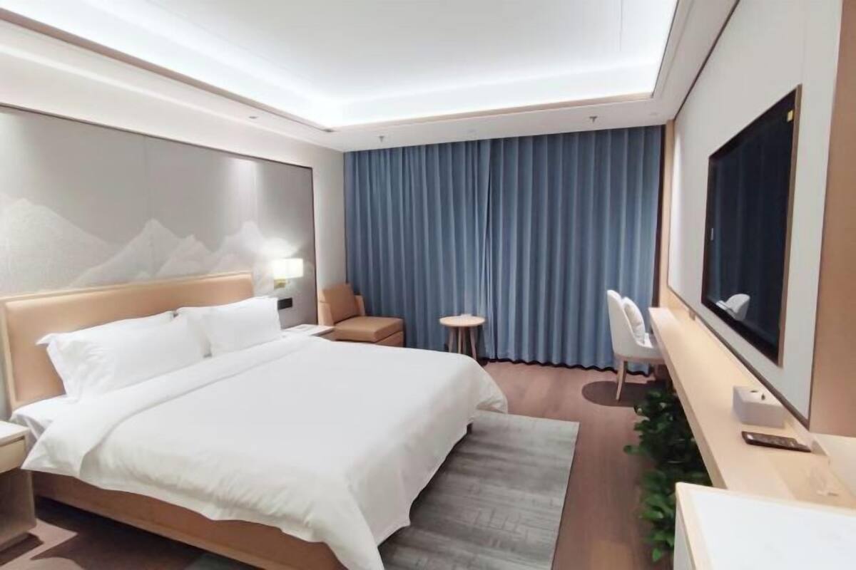 A modern hotel room with a large bed, a desk, a TV, and a view of the mountains through the curtains.