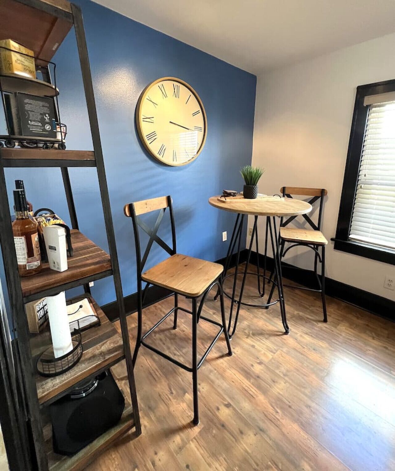 A room with a blue wall, a round wall clock, a wooden table with two chairs, a shelf with various items, and a window with blinds.