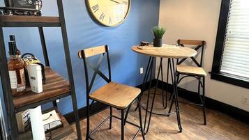 A room with a blue wall, a round wall clock, a wooden table with two chairs, a shelf with various items, and a window with blinds.