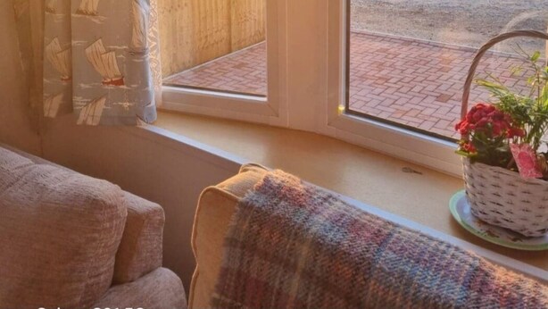 Una habitación con un ventanal grande, vista al atardecer sobre un campo, y una cesta de mimbre con flores en el alféizar.