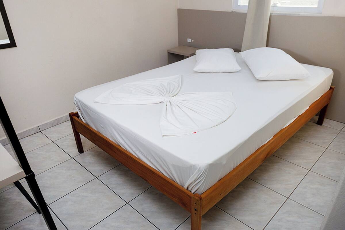 A neatly made bed with white linens and a wooden frame, a small bedside table, and a window with curtains.