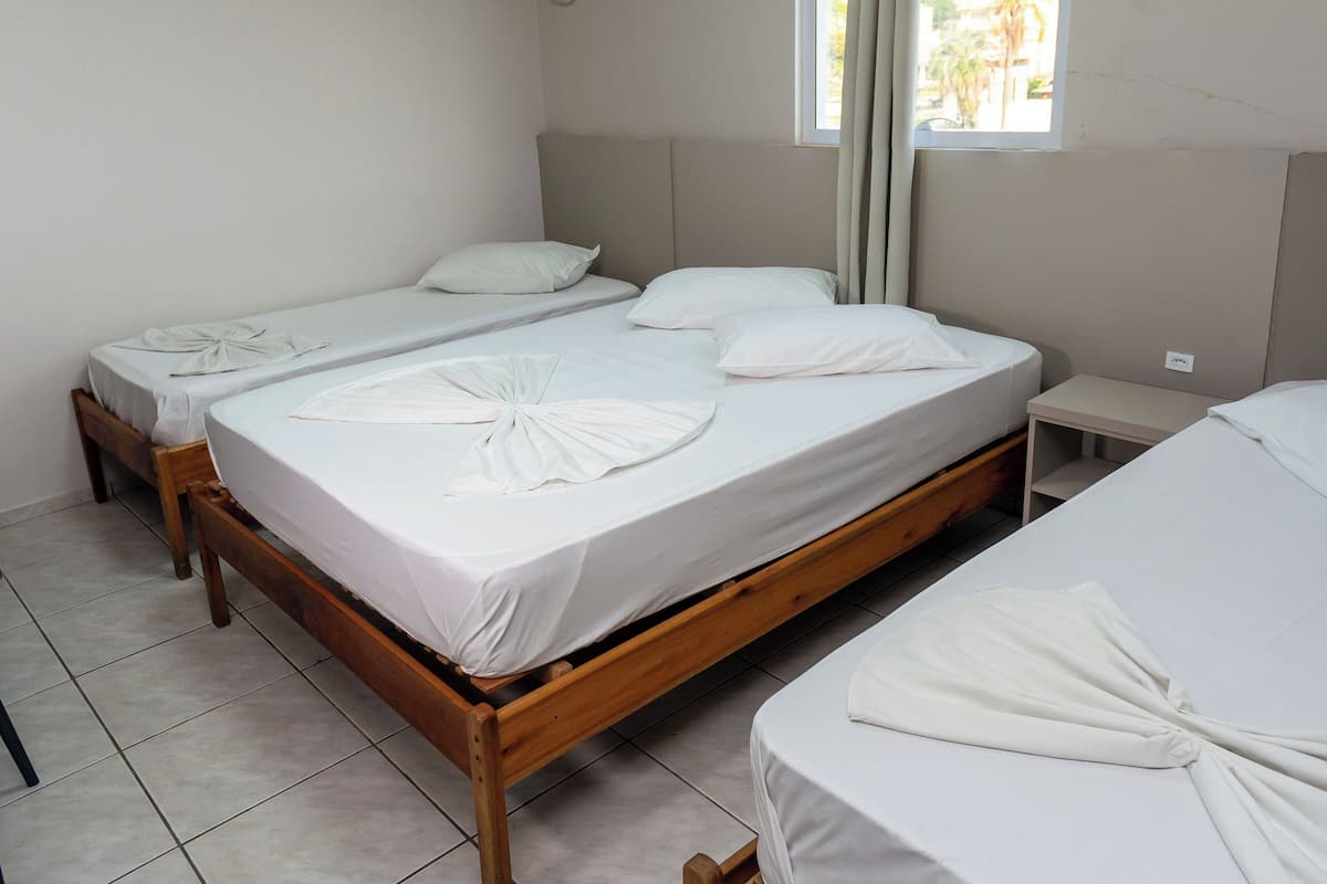 A hotel room with three single beds, a ceiling light, an air conditioning unit, and a window with curtains.
