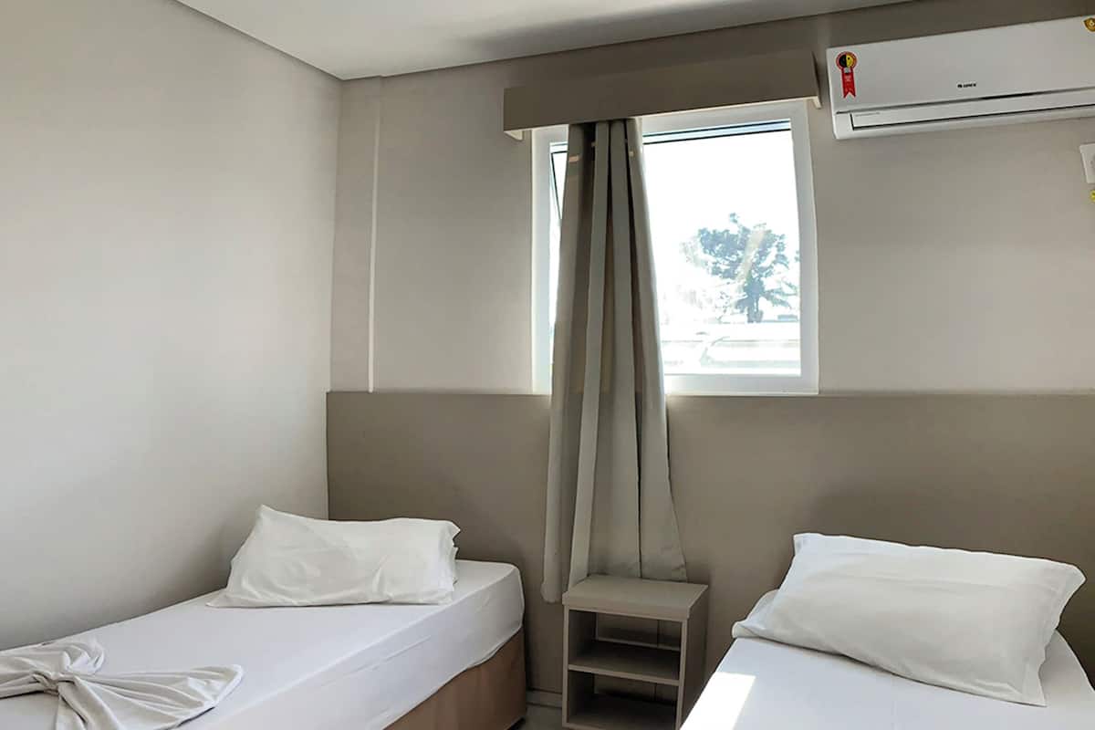 A hotel room with two single beds, a small table, a window with curtains, and an air conditioning unit.