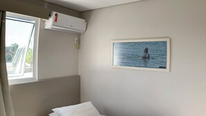 A single bed with a neatly made white sheet and a decorative knot on the side, a small bedside table, a window with a view of trees, and an air conditioning unit on the wall. - Ok Inn Hotel Tubarão (Tubarao)