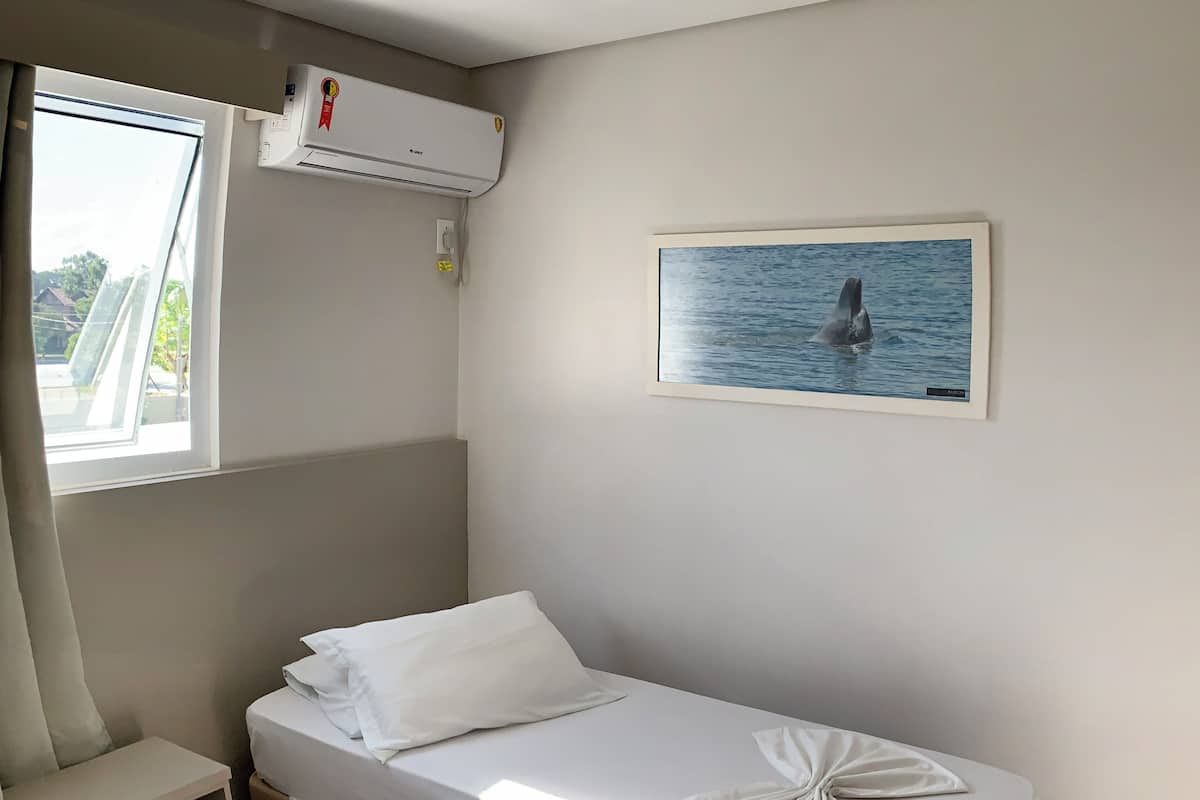 A single bed with a neatly made white sheet and a decorative knot on the side, a small bedside table, a window with a view of trees, and an air conditioning unit on the wall.