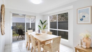 Un comedor con una mesa de madera y sillas de mimbre, ventanas amplias y una planta en maceta.