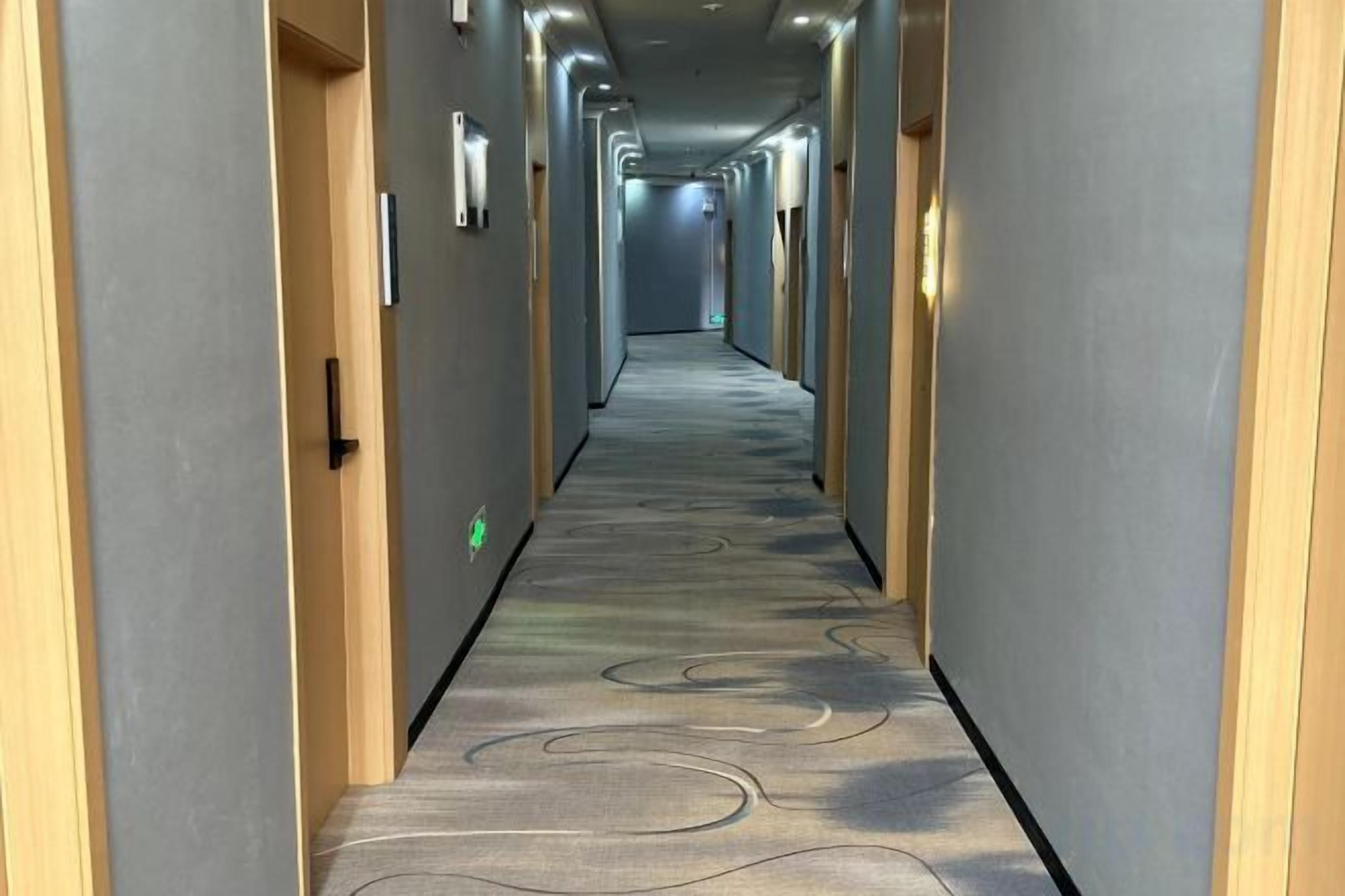 Un couloir d’hôtel avec un sol recouvert de moquette, des portes de chaque côté et un éclairage au plafond.