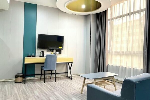 Una habitación de hotel moderna con piso de madera, un escritorio con televisor de pantalla plana, una silla azul, una mesita y un ventanal grande con cortinas.