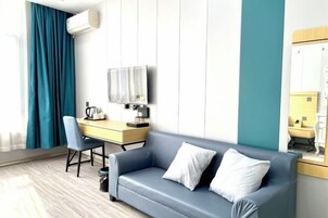 Quarto de hotel com um sofá azul, uma escrivaninha com cadeira, televisão e um espelho grande.