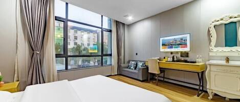 A hotel room with a large bed, a desk with a computer, a TV, and a view of buildings outside the window.