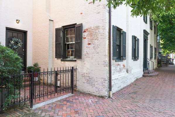 A brick-paved alleyway with a white building featuring a black door and shuttered windows. - Spacious Converted Carriage House in Historic District near USNA (Annapolis)