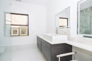 A modern bathroom with a large white sink, dark cabinetry, and a large mirror. There is a window with a brick wall view and framed pictures on the wall. - Spacious Converted Carriage House in Historic District near USNA (Annapolis)