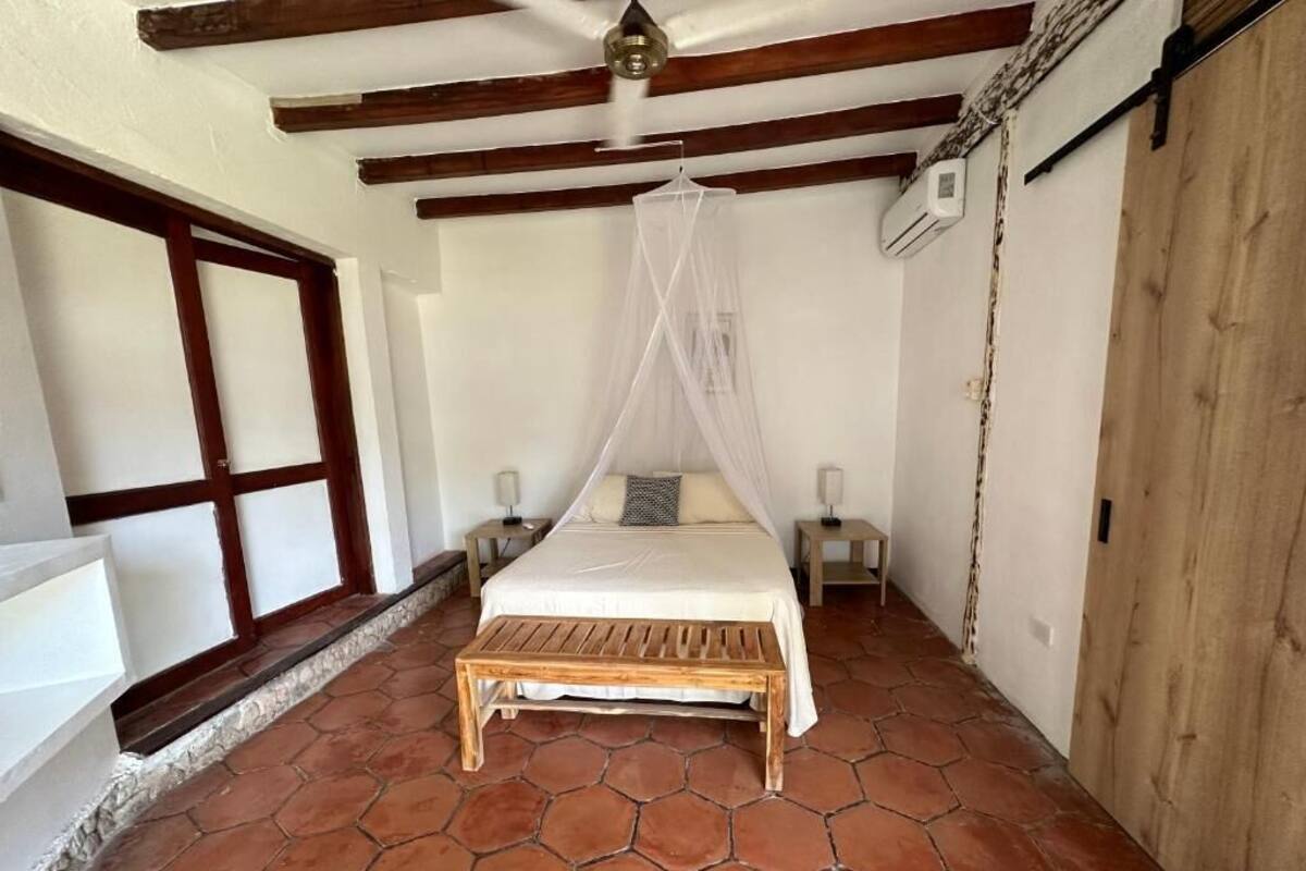 A bedroom with a wooden bed, a bench, a canopy, and a stone-tiled floor.