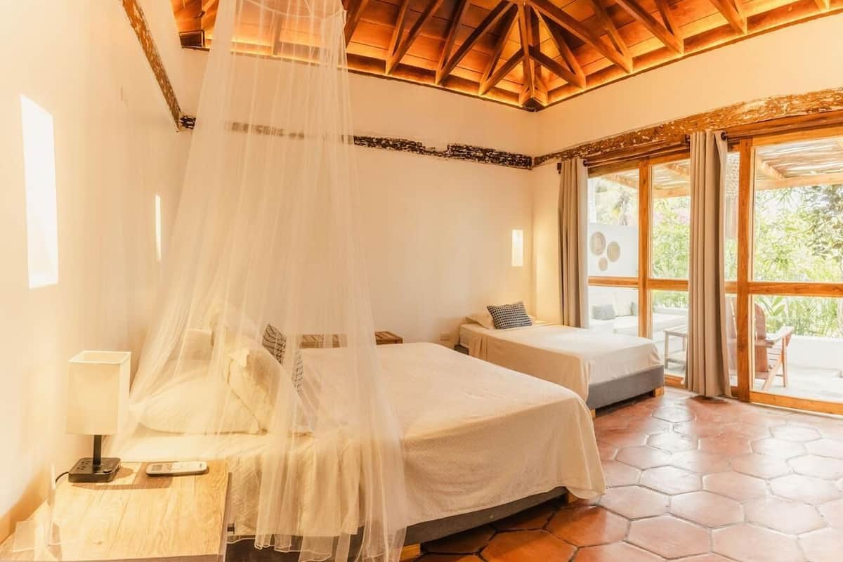 A bedroom with two beds, a canopy bed, a wooden ceiling, and a large window with curtains.