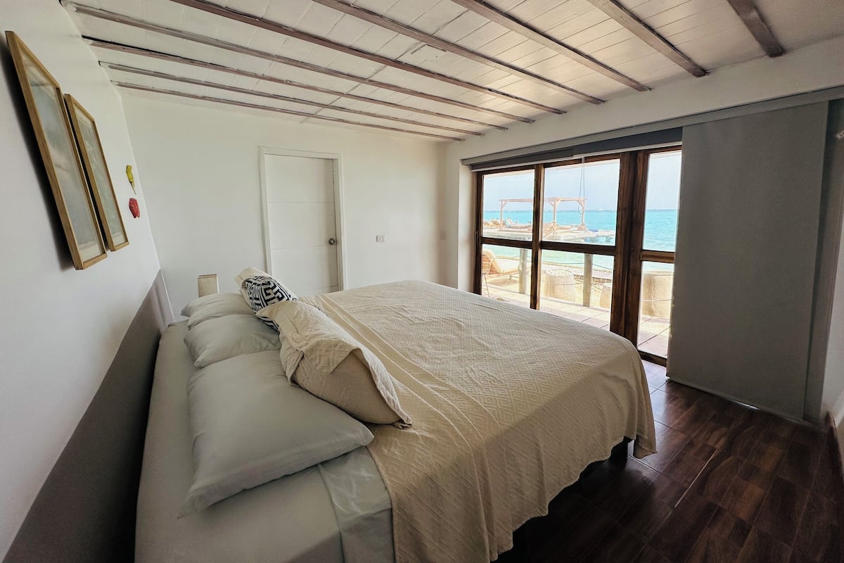A bedroom with a large bed, a door, and a window with a view of the sea.