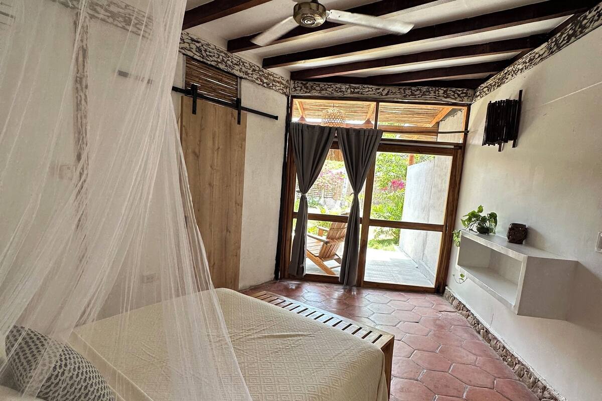 A bedroom with a bed under a canopy, a ceiling fan, a view of the outdoors through a window, and a tiled floor.