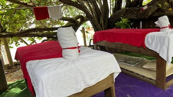 Guests can relax under the shade of a large tree, surrounded by a rustic outdoor setup featuring two beds with red and white bedspreads.