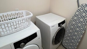 Un área de lavandería con dos lavadoras de carga frontal blancas, una cesta blanca para ropa y un estante con productos de limpieza.
