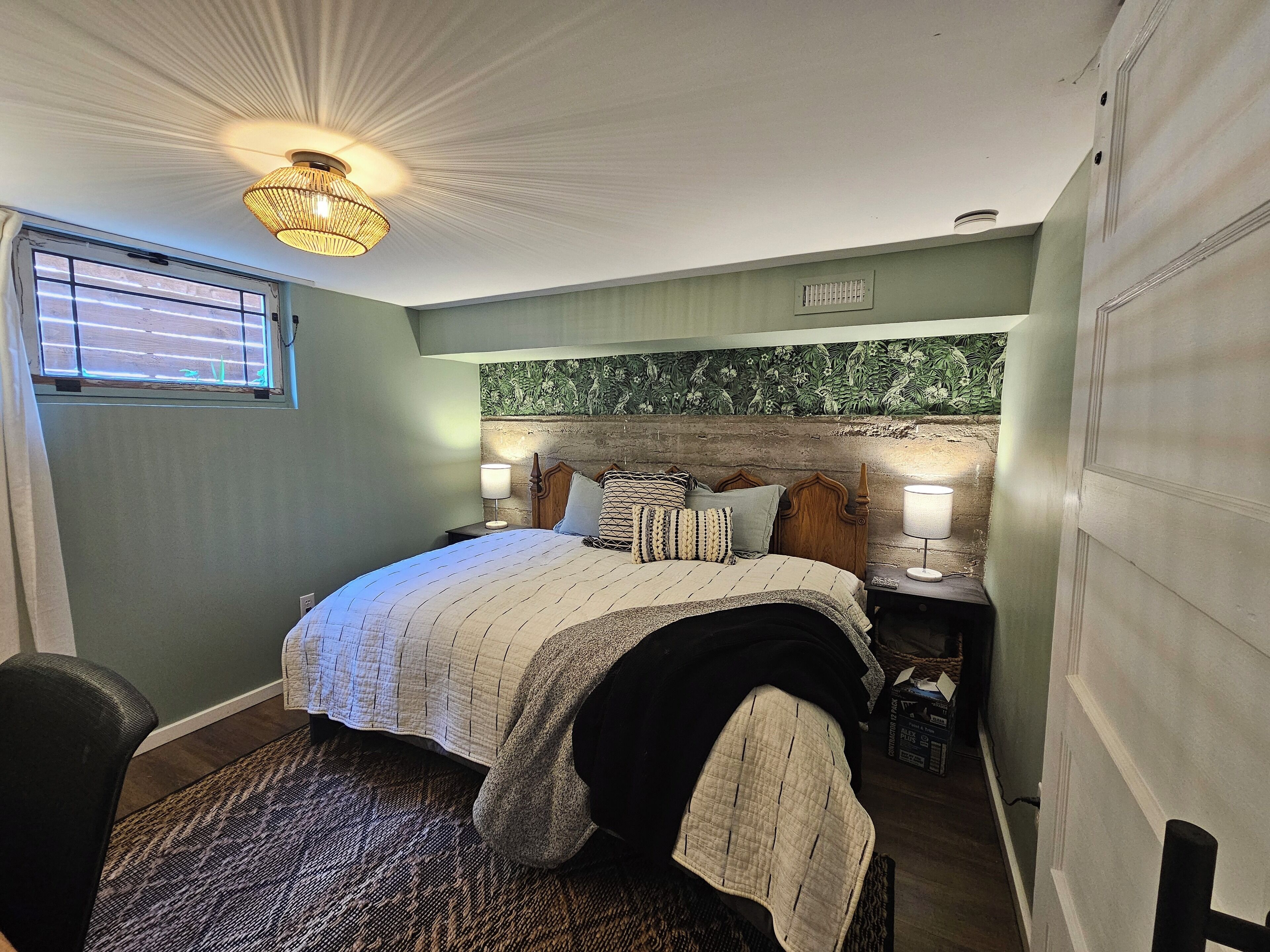 A bedroom with a bed, two bedside tables, a wall-mounted light fixture, a window with blinds, and a textured wall.