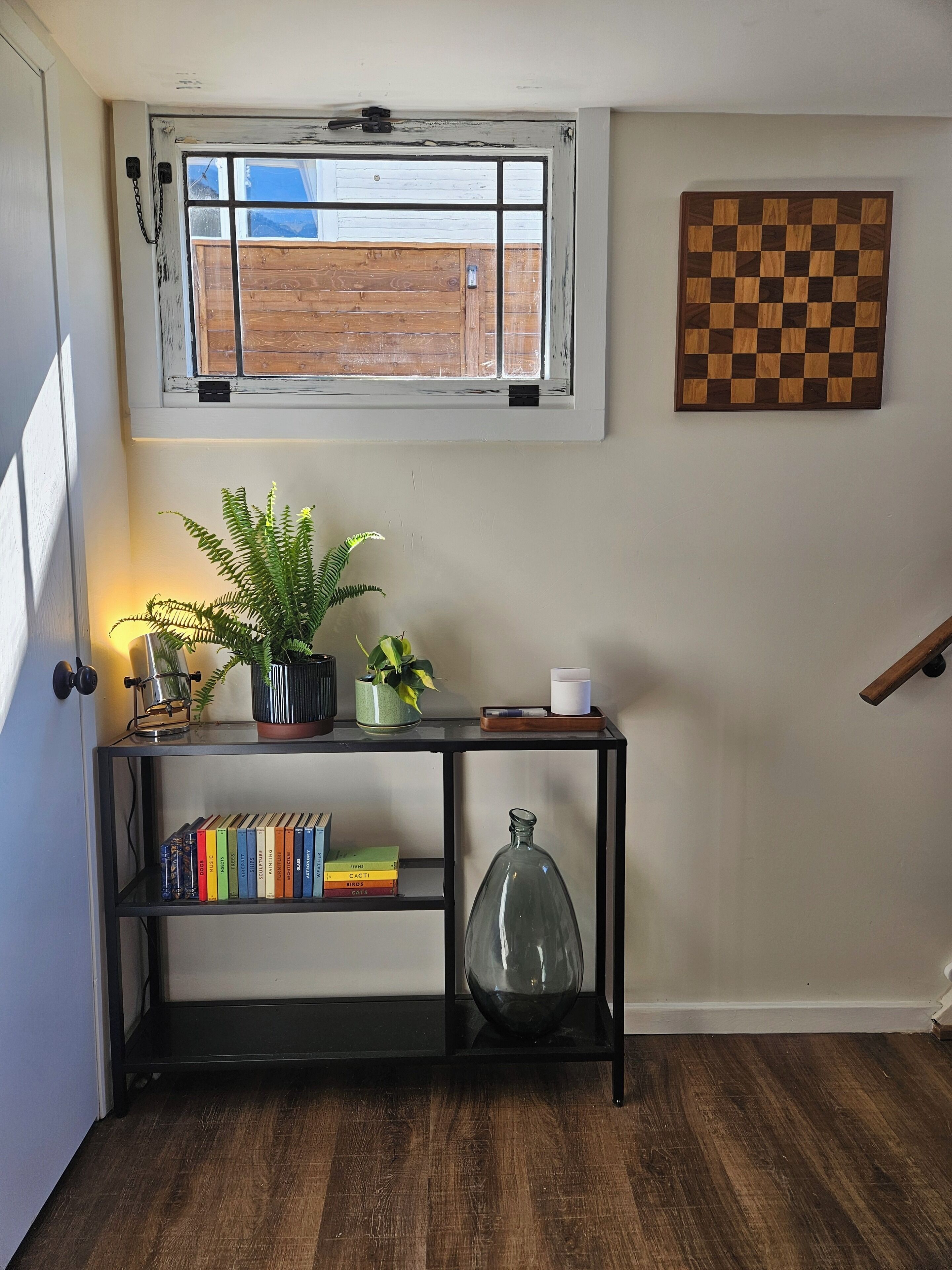 A room with a wooden floor, a window with a view of a wooden fence, a black metal shelf with books, a potted plant, and a glass vase.
