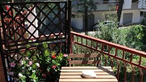 Une table en bois avec un coussin, placée sur un balcon doté d’une rambarde rouge, et entourée de plantes et de fleurs en pot.