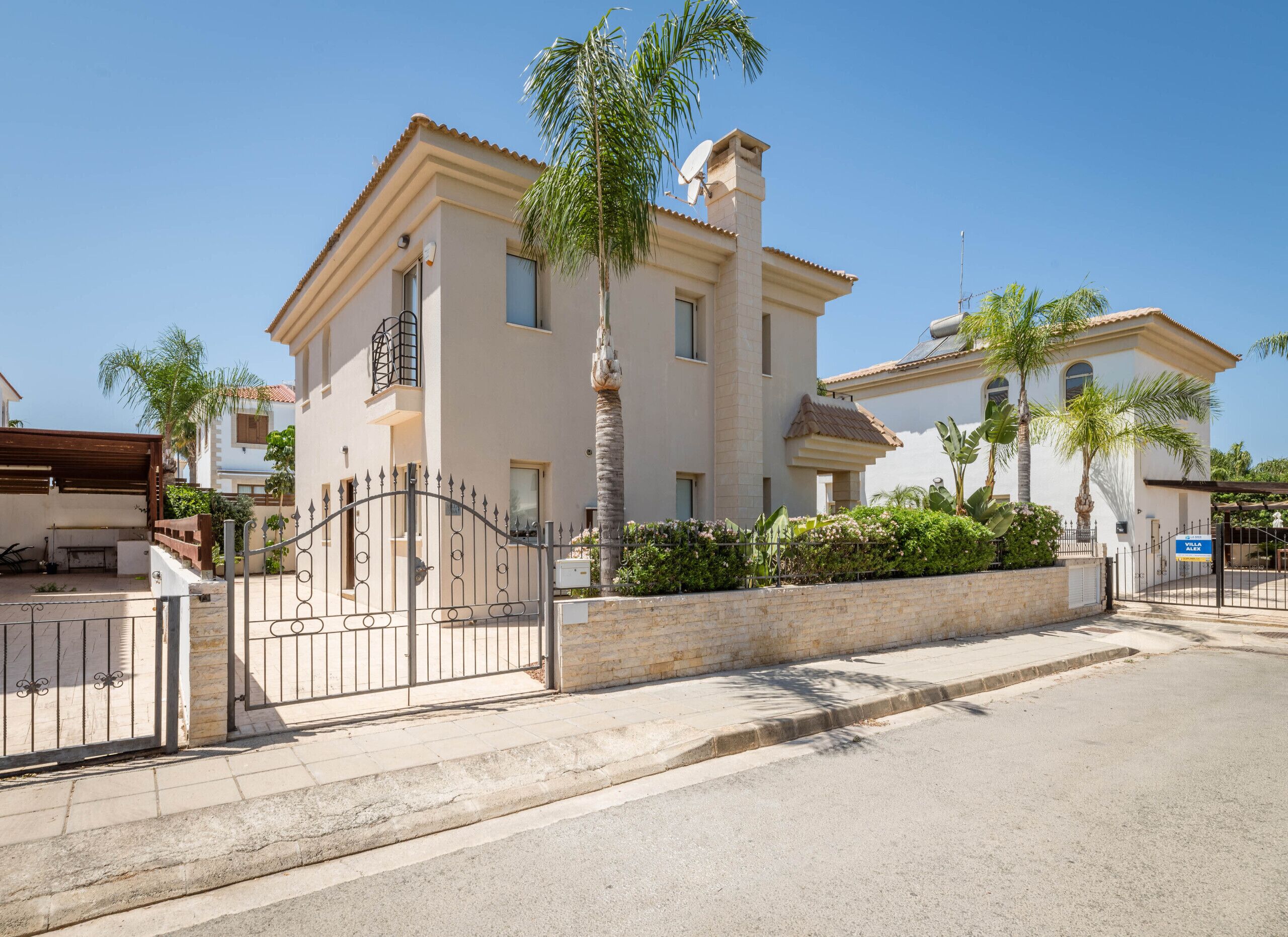 A two-story building with a gated entrance, a stone wall, and palm trees.