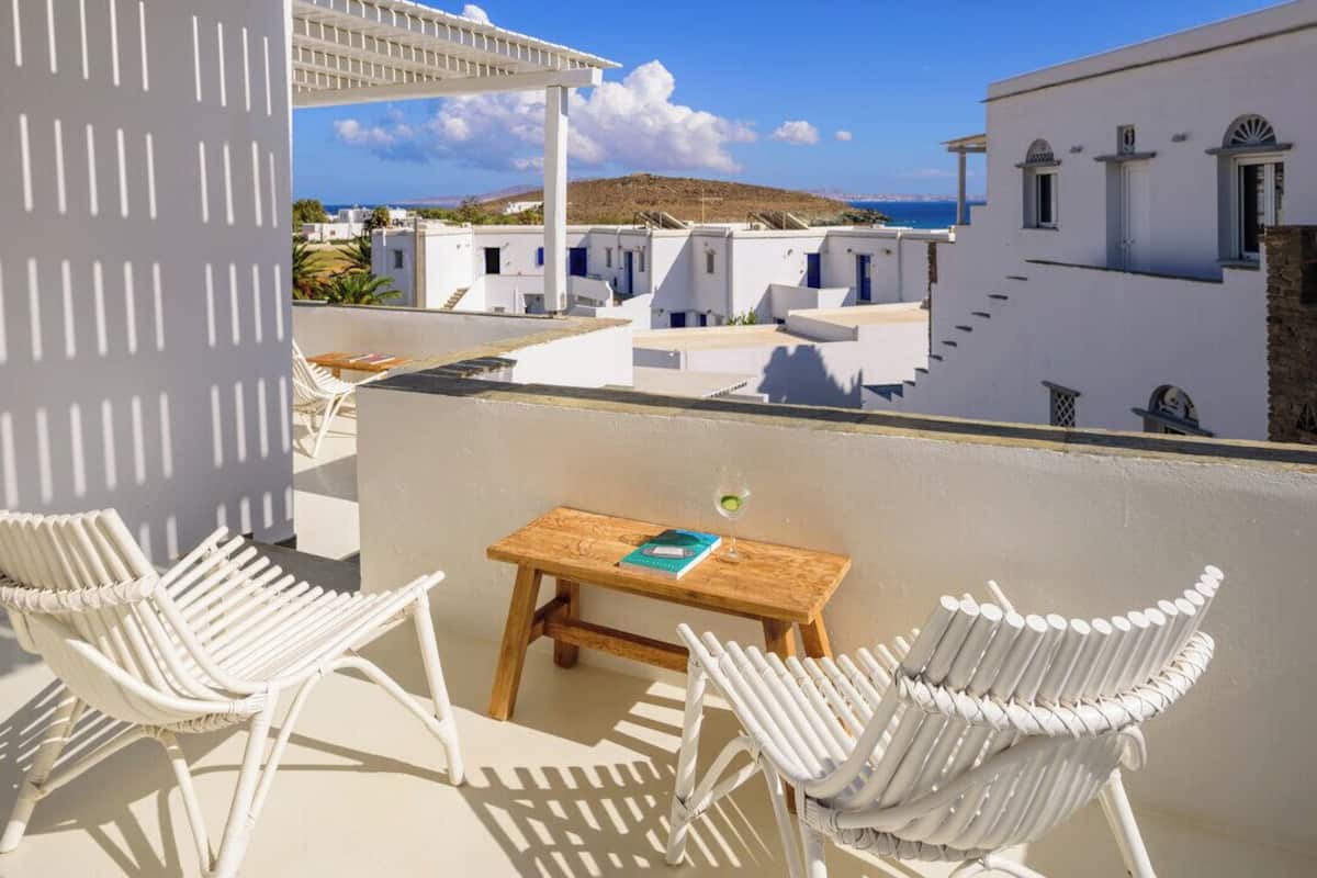 Une terrasse dotée de chaises en osier blanc et d’une table en bois, offrant une vue sur un village côtier aux bâtiments blancs et sous un ciel d’un bleu limpide.