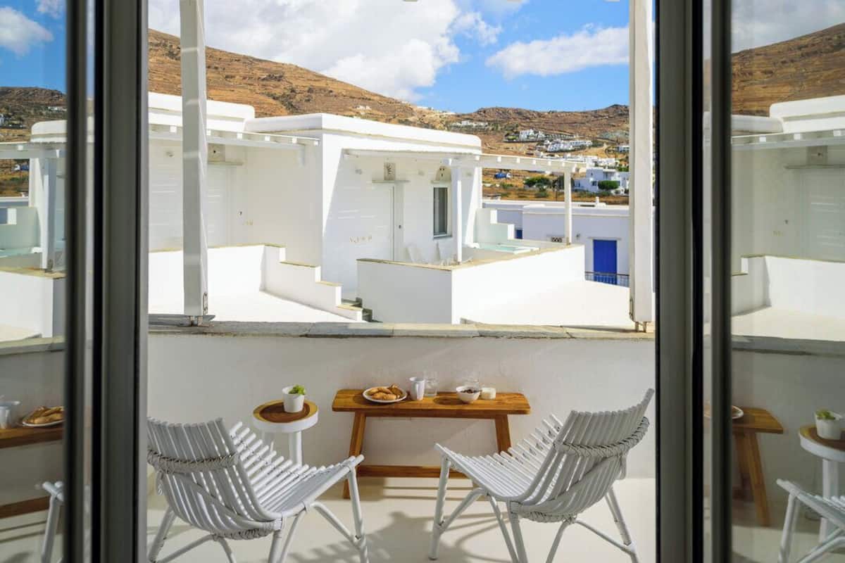 Un balcon agrémenté de chaises en osier blanc et d’une petite table, d’où l’on peut admirer un ensemble d’immeubles blancs, avec une chaîne de montagnes en toile de fond.