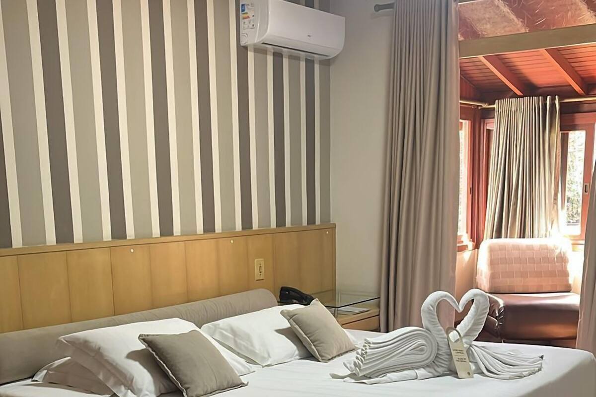 Habitación de hotel con una cama grande, cabecera, unidad de aire acondicionado, suelo de madera y ventana con cortinas.