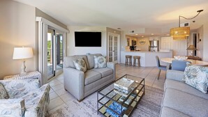 A living room with a sofa, coffee table, and a TV. There is a kitchen area with a dining table and chairs. - End Unit; Ocean Peek; 5th floor - Villamare. 2 hrs Pickleball/Tennis per day! (Hilton Head Island)