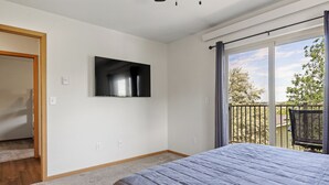 Un dormitorio con una cama grande, un televisor de pantalla plana montado en la pared y un balcón con vistas a árboles y un edificio.