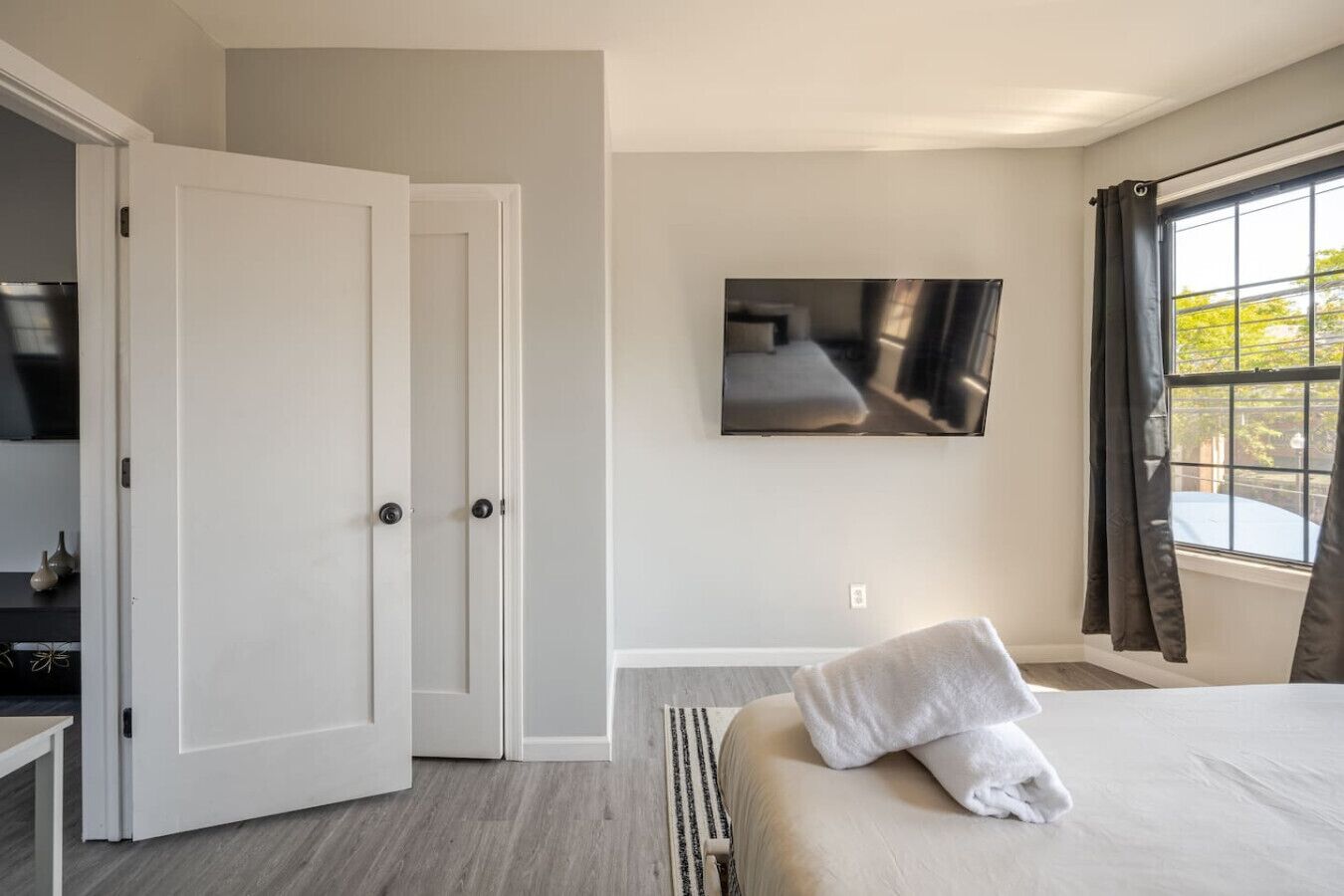 Ein Schlafzimmer mit einem Bett, einem an der Wand montierten Fernseher, einem Fenster mit Vorhängen und einer Tür.
