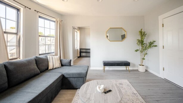 A modern living room with a sectional sofa, a bench, a coffee table, a potted plant, and a large mirror on the wall. - Modern 3-Bdrm Apt in Black Rock (Bridgeport)