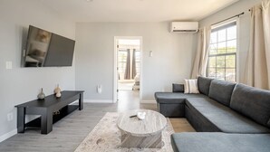 A modern living room with a sectional sofa, a coffee table, a TV mounted on the wall, and an air conditioning unit. - Modern 3-Bdrm Apt in Black Rock (Bridgeport)