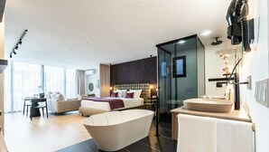 Una habitación de hotel moderna con una bañera grande, una zona de estar separada con sofá y una mesa de comedor, un área de ducha con paredes de vidrio y un ventanal que proporciona luz natural.