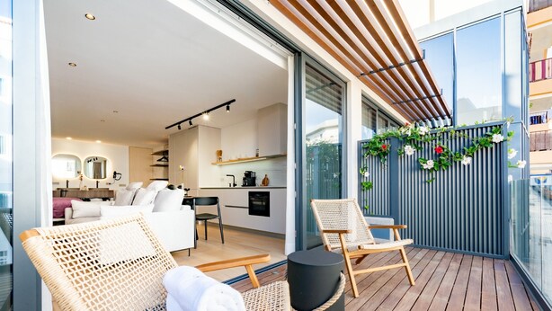 Un balcón con sillas de mimbre, un reposapiés negro redondo y una puerta de cristal que da a una cocina y sala de estar modernas.